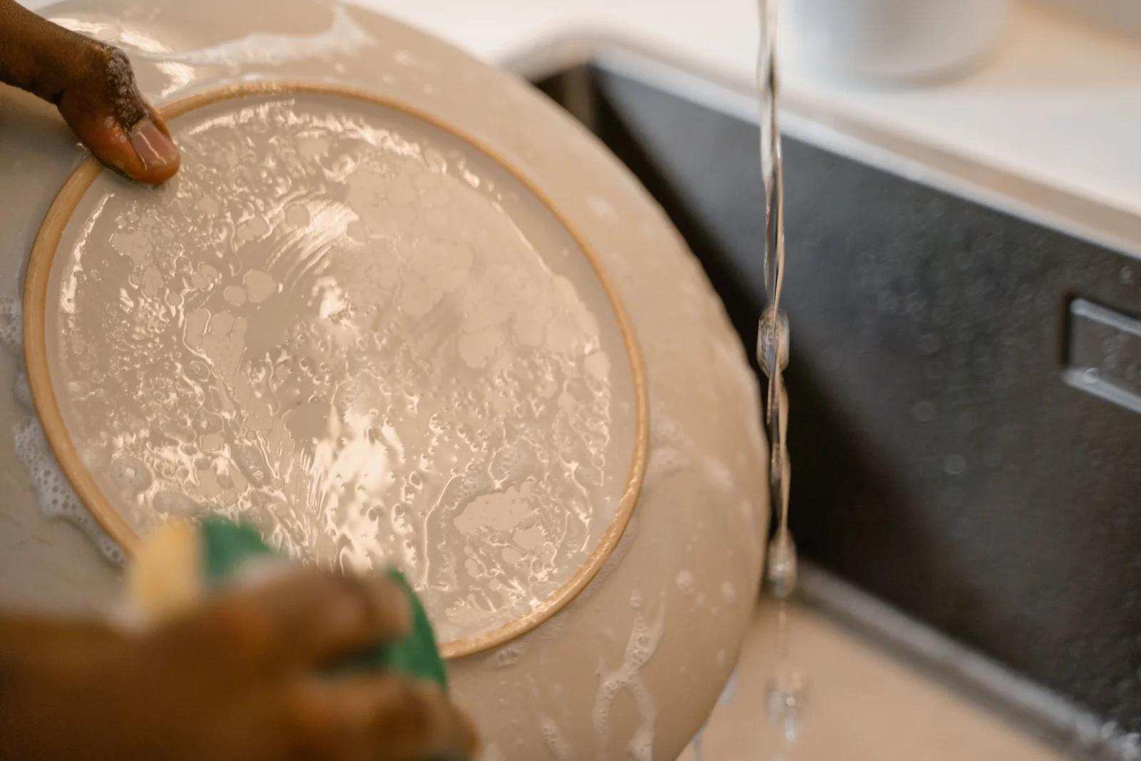 Hands wash a ceramic plate with a sponge under running water.