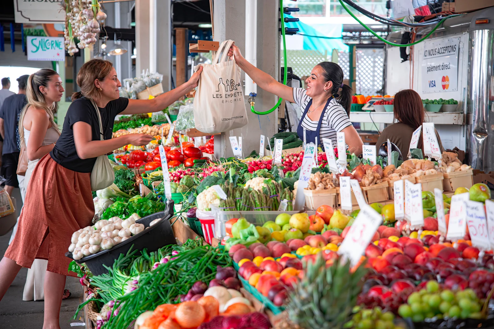 A vendor hands a shopping bag to a customer across a colourful produce stall.
